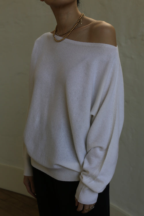 A person wearing an off-the-shoulder, relaxed-fit, Ballerina's Cashmere Sweater | Ivory paired with black pants. They are accessorized with a gold chain necklace and several rings. The background is a soft, neutral-colored wall. The person's head is not visible in the image.