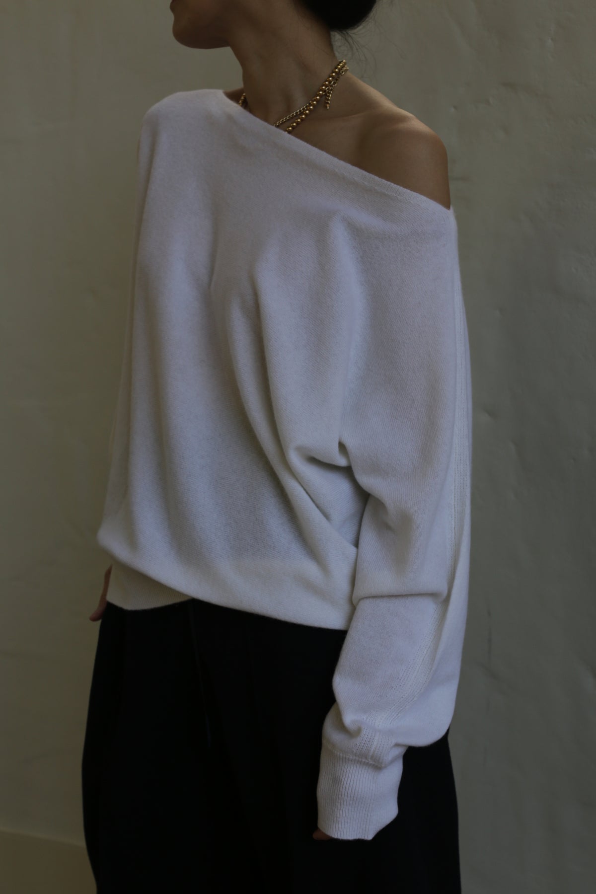 A person wearing an off-shoulder, relaxed fit Ballerina's Cashmere Sweater | Ivory and dark trousers stands against a plain wall. They have a gold necklace around their neck, and their head is turned to the side, obscuring their face.