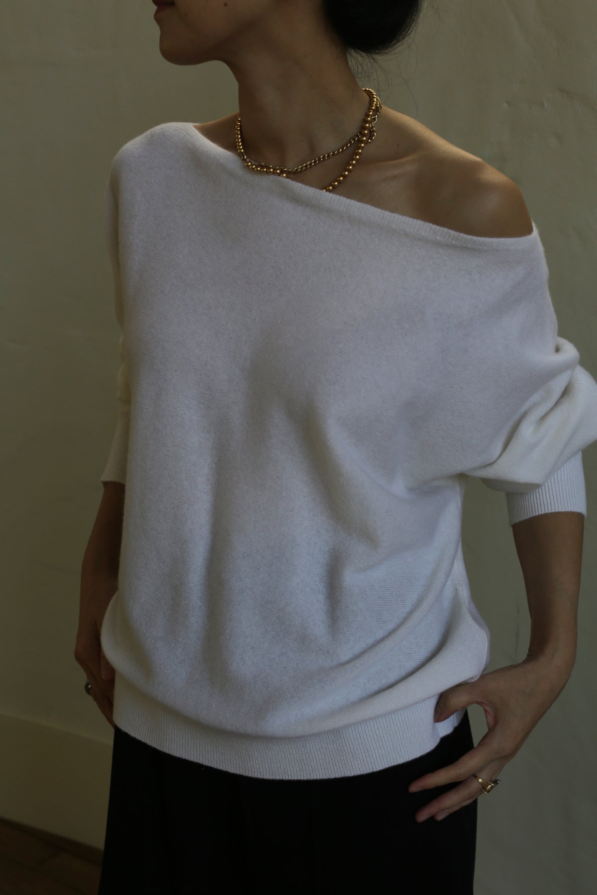 A person wearing a white, relaxed fit, off-the-shoulder Ballerina's Cashmere Sweater | Ivory and a gold necklace stands against a plain background. Their hands rest on their hips, and their face is not visible.