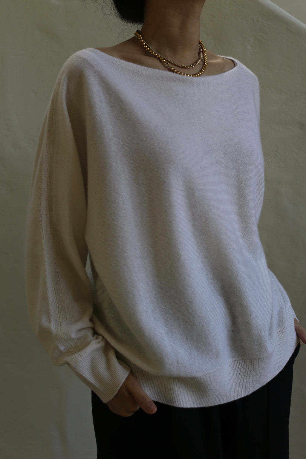 A person wearing a relaxed fit, light-colored Ballerina's Cashmere Sweater | Ivory and black pants stands against a plain, light background. They have their hands in their pockets and are accessorized with a gold chain necklace. Their face is not visible.