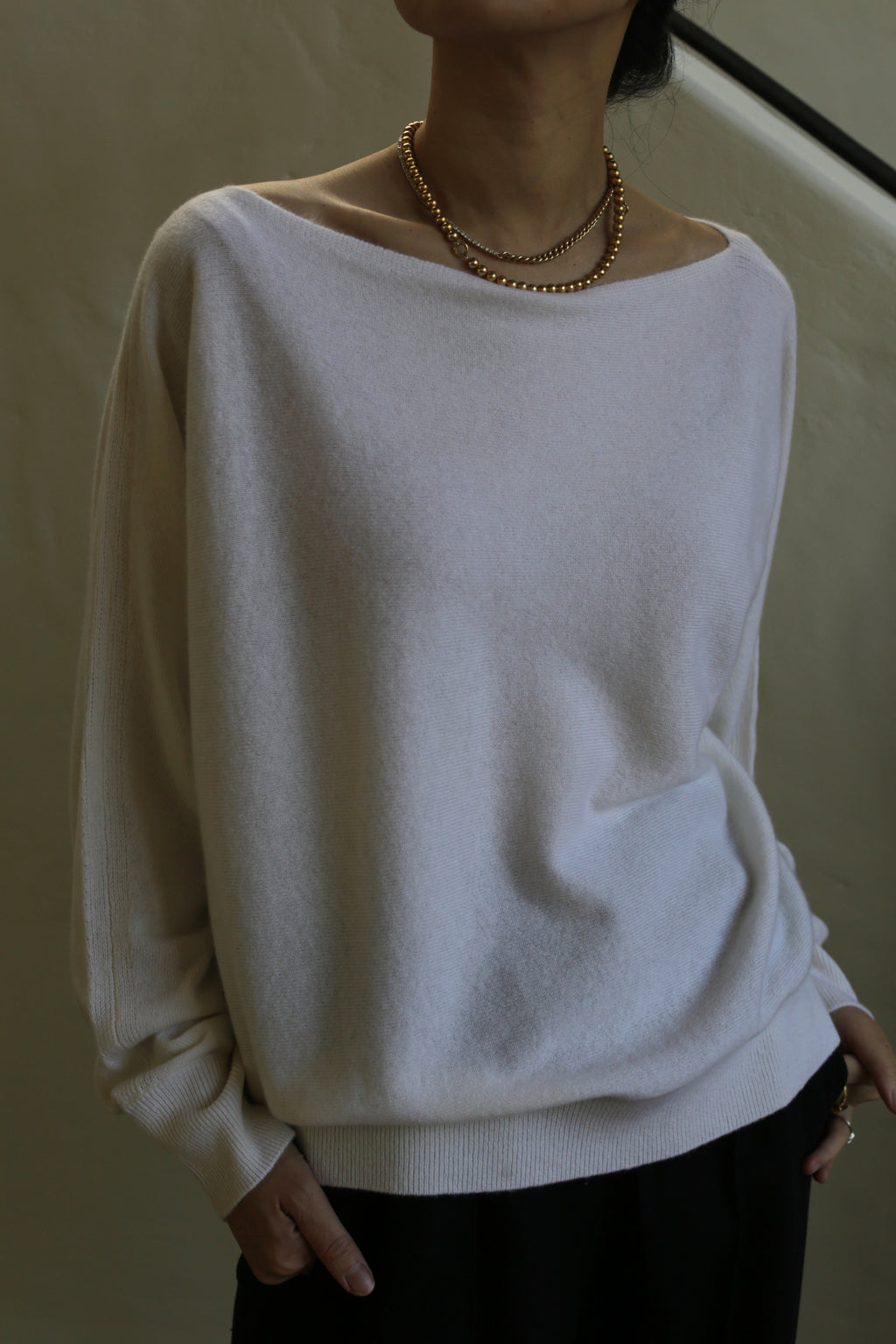 A person wearing a loose, light-colored Ballerina's Cashmere Sweater | Ivory with a boat neck and a simple, elegant necklace stands against a neutral background. The sweater has a relaxed fit and the person's hands are casually placed in their pockets.