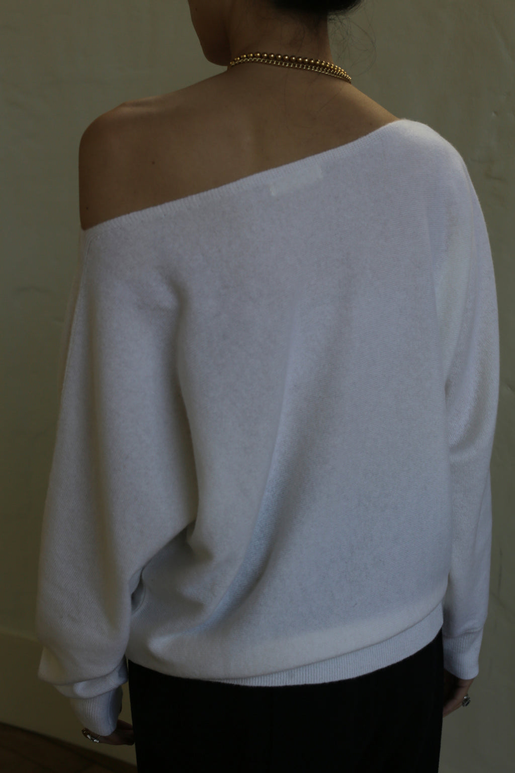 A person is seen from the back wearing a relaxed fit, white Ballerina's Cashmere Sweater | Ivory with an exposed shoulder. They also have a gold chain necklace around their neck and dark pants. The background is plain and unadorned.