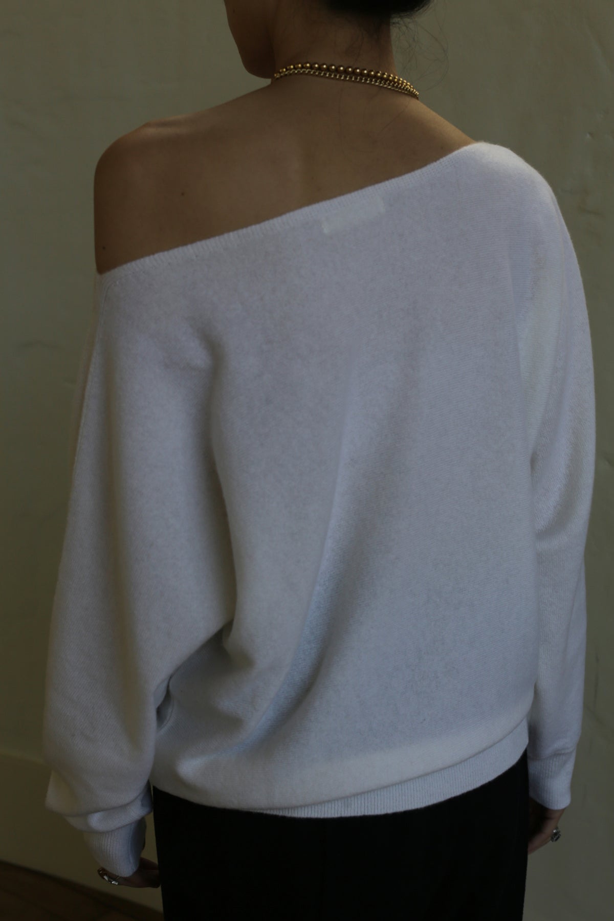 A person is seen from the back wearing a relaxed fit, white Ballerina's Cashmere Sweater | Ivory with an exposed shoulder. They also have a gold chain necklace around their neck and dark pants. The background is plain and unadorned.