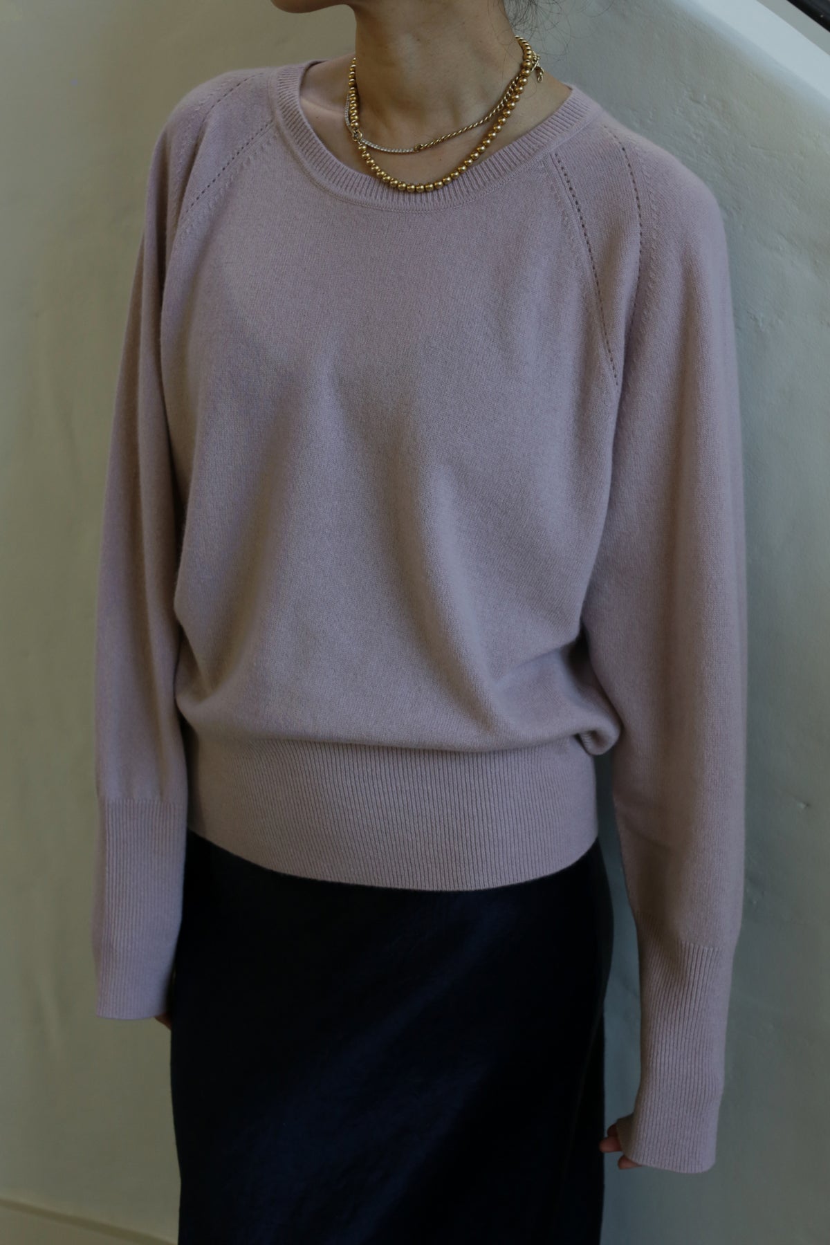 A person is wearing a light purple Raglan Crewneck Cashmere Sweater | Peony with long sleeves and a black skirt. They have a double-layered gold necklace around their neck. The background is a plain, light-colored wall. This cozy crewneck ensemble is the perfect wardrobe staple for any season.
