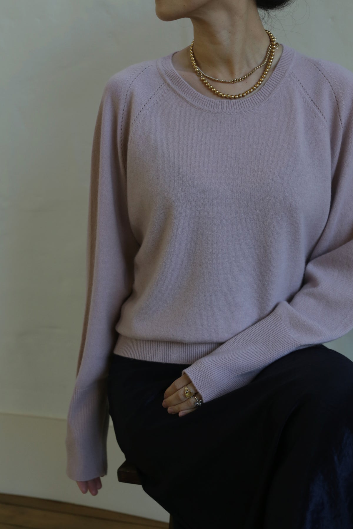 A person wearing a Raglan Crewneck Cashmere Sweater | Peony and a dark skirt, accessorized with gold necklaces and rings, captured from the neck down. The person's left hand rests on their lap against a neutral background, showcasing this sleek wardrobe staple.