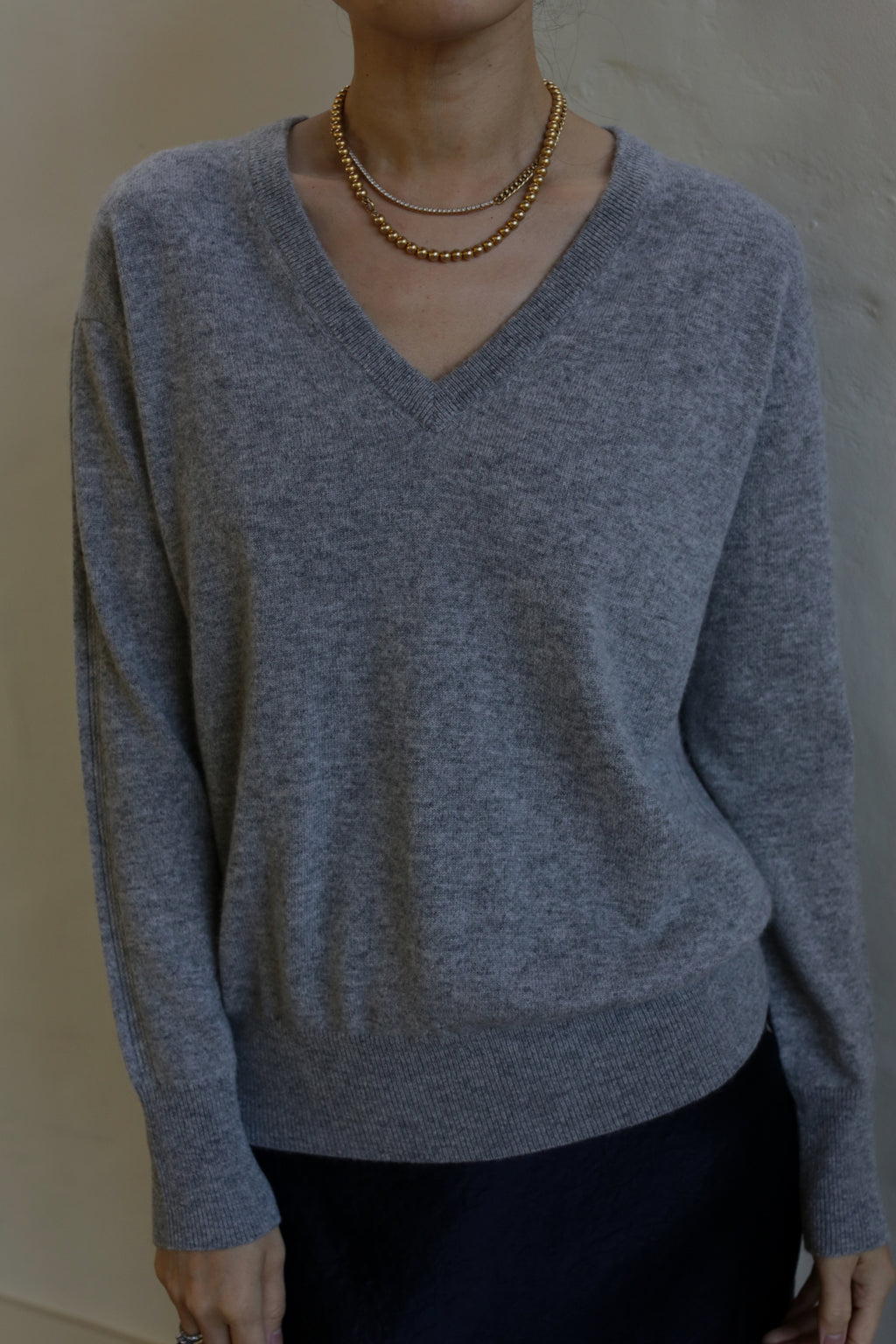 Person wearing a light gray V-Neck Cashmere Sweater | Grey, layered gold necklaces, and a dark skirt, standing against a plain background. The 100% cashmere outfit is casual and stylish. The person's face is not visible in the image.