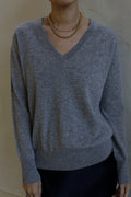 Person wearing a light gray V-Neck Cashmere Sweater | Grey, layered gold necklaces, and a dark skirt, standing against a plain background. The 100% cashmere outfit is casual and stylish. The person's face is not visible in the image.
