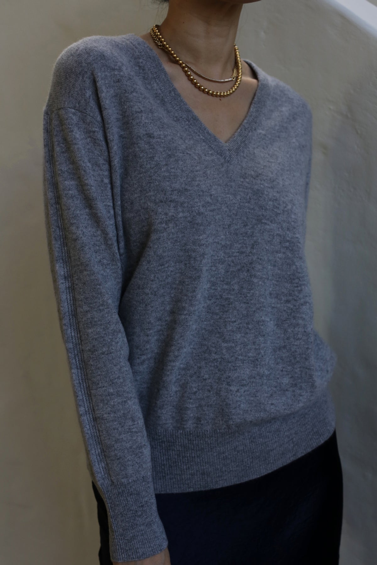 Person wearing a light grey V-Neck Cashmere Sweater | Grey and a layered gold necklace against a plain background. The image captures the upper body and focuses on the outfit details, highlighting the relaxed fit of the 100% cashmere garment.
