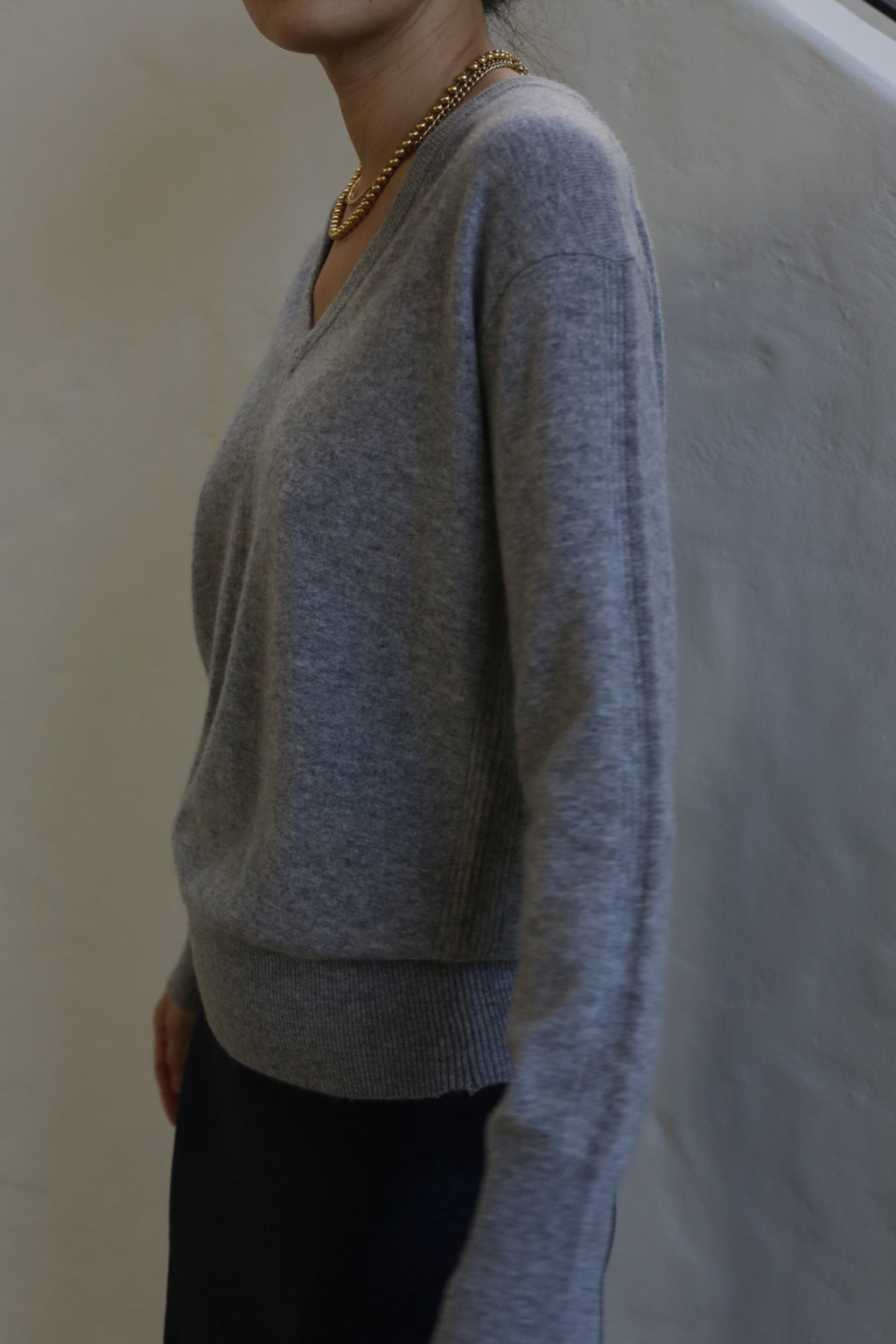 A person wearing a relaxed fit, V-Neck Cashmere Sweater | Grey and a gold chain necklace stands against a neutral, light-colored background. The person is partially out of frame, showing only their torso and part of their face.
