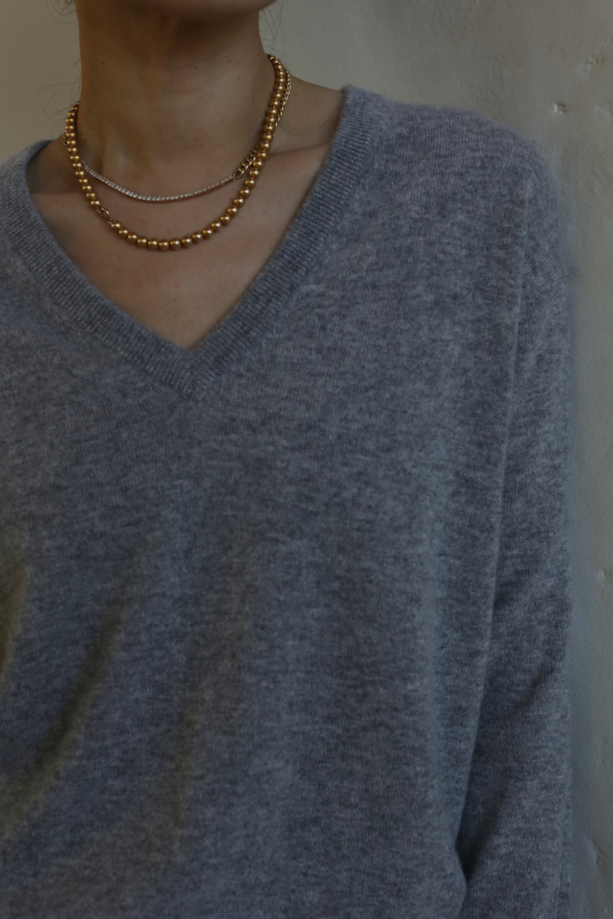 A person is wearing a light grey, relaxed fit V-Neck Cashmere Sweater | Grey and two layered necklaces: one with small spherical beads and the other with tiny beads. The background is a plain wall, and the photo is cropped to show the person's upper torso and part of their neck.
