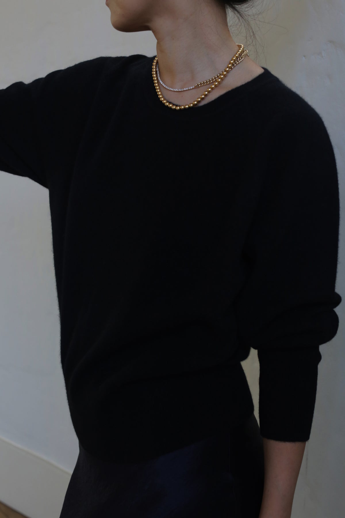 A person with their hair tied back is wearing a medium-weight Raglan Crewneck Cashmere Sweater | Black and two layered gold necklaces. They are standing against a plain, light-colored background with their face turned away from the camera, displaying part of their neck and shoulder.