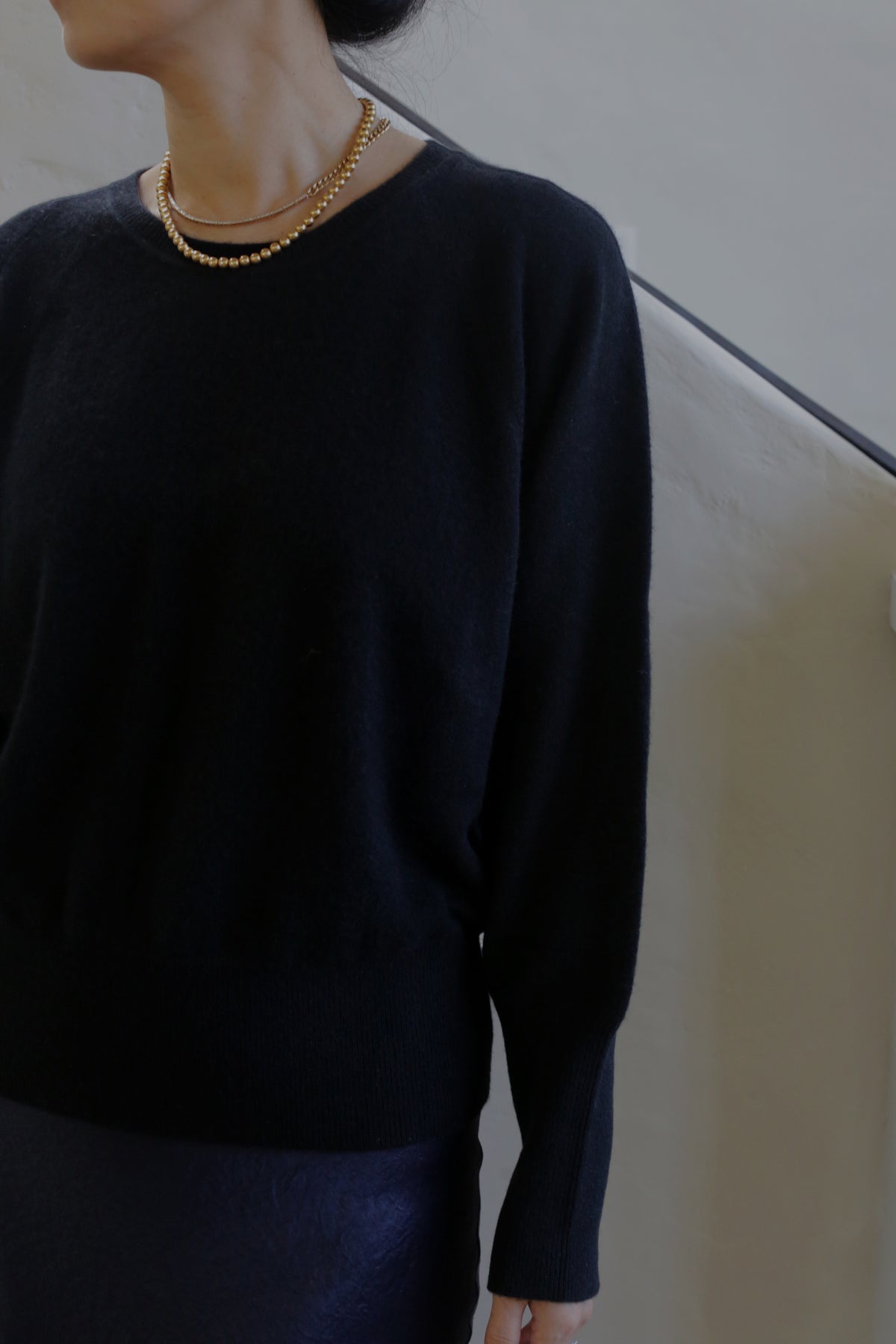 A person with dark hair is shown from the shoulders up, wearing a Raglan Crewneck Cashmere Sweater | Black and a gold beaded necklace. The background appears to be a plain, light-colored wall. The individual's facial features are not visible.