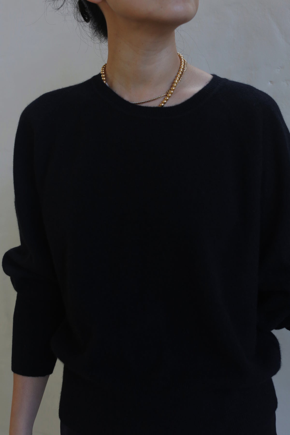 A person is wearing a black, medium-weight, long-sleeve Raglan Crewneck Cashmere Sweater | Black with a simple gold necklace. They are standing against a neutral background with their head tilted up and slightly to the side, revealing only part of their face.