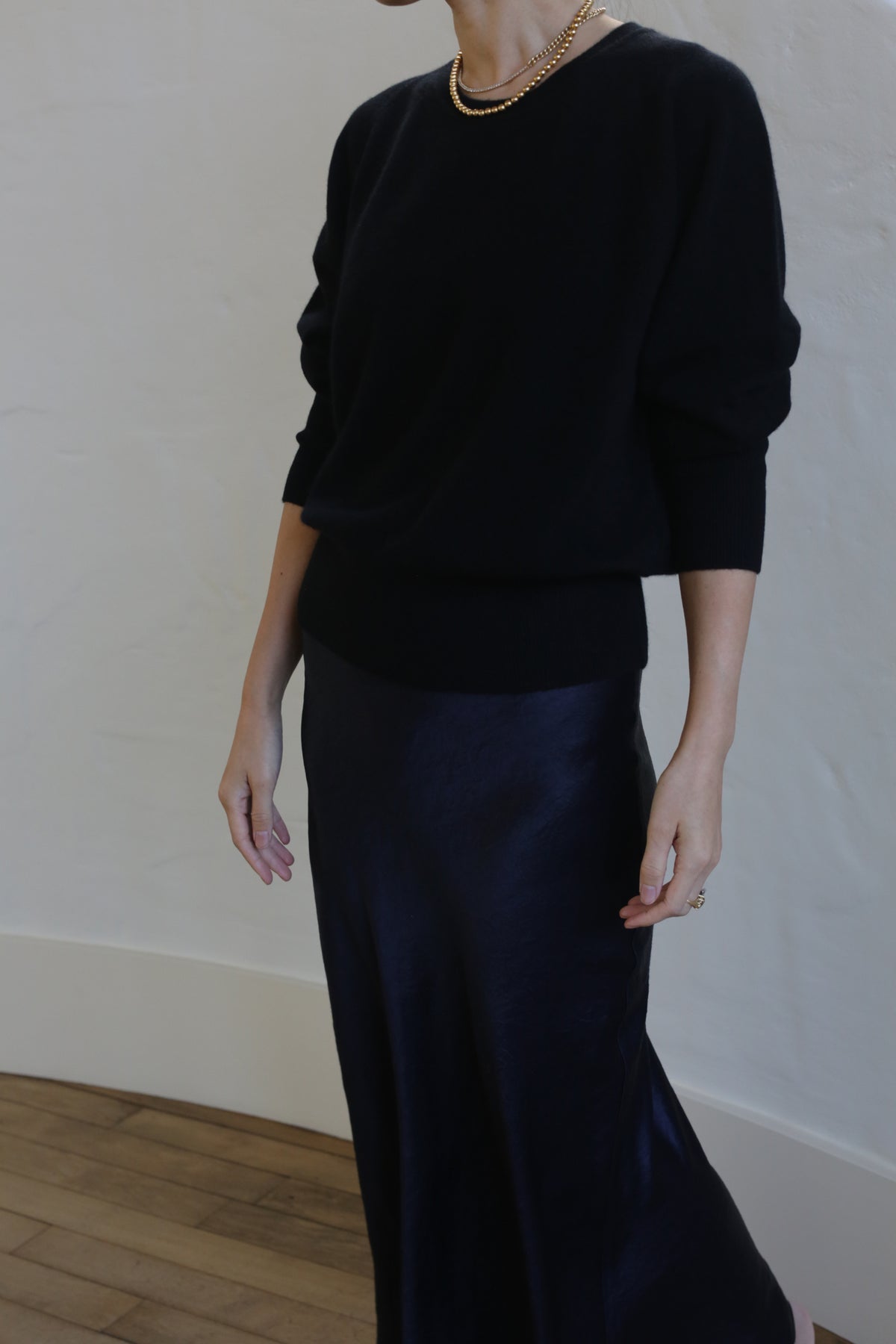 A person wearing a black long-sleeve top and a Liquid Slip Dress Long | Midnight stands against a neutral background. They have a gold necklace around their neck and a ring on their right hand. The image shows their upper body and part of their lower body, but not their face.