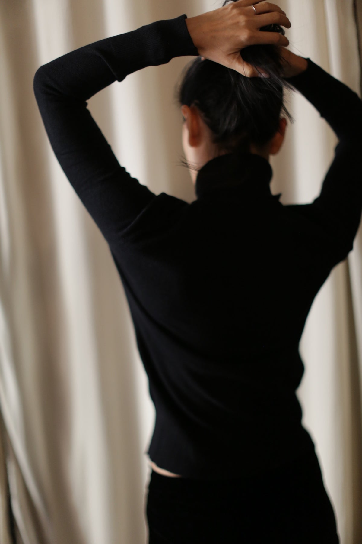 A person stands with their back to the camera, facing a light-colored curtain. They are wearing a black long-sleeved Cashmere Turtleneck and have their arms raised, hands gathered at the back of their head as if fixing their hair. The background is softly blurred.