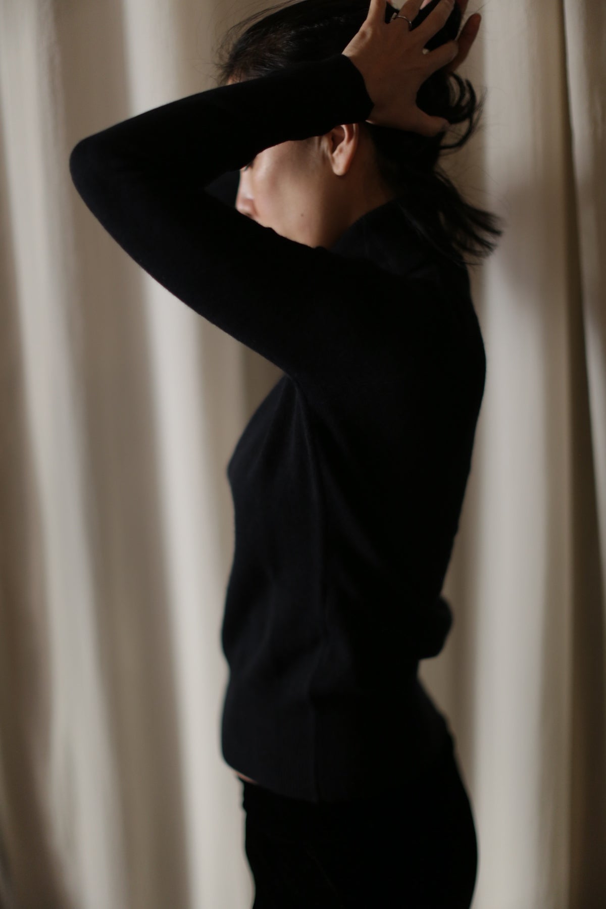 A person with dark hair is standing in profile, facing left, and adjusting their hair with both hands. They are wearing a black, long-sleeve Cashmere Turtleneck. The background features white, flowing curtains.