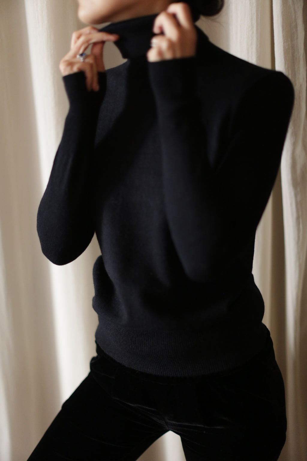 A person wearing a black, hand washable, long-sleeve **Cashmere Turtleneck** stands in front of a light-colored curtain. The individual's face is obscured, and they are seen adjusting the collar of the turtleneck with both hands.