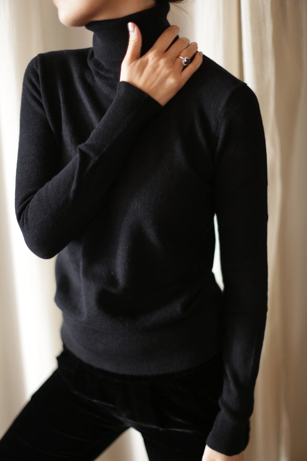 Person wearing a black Cashmere Turtleneck and black pants, standing against a light-colored curtain backdrop. One hand is touching the collar of the Cashmere Turtleneck, and a ring is visible on one of the fingers. The face is mostly out of the frame.