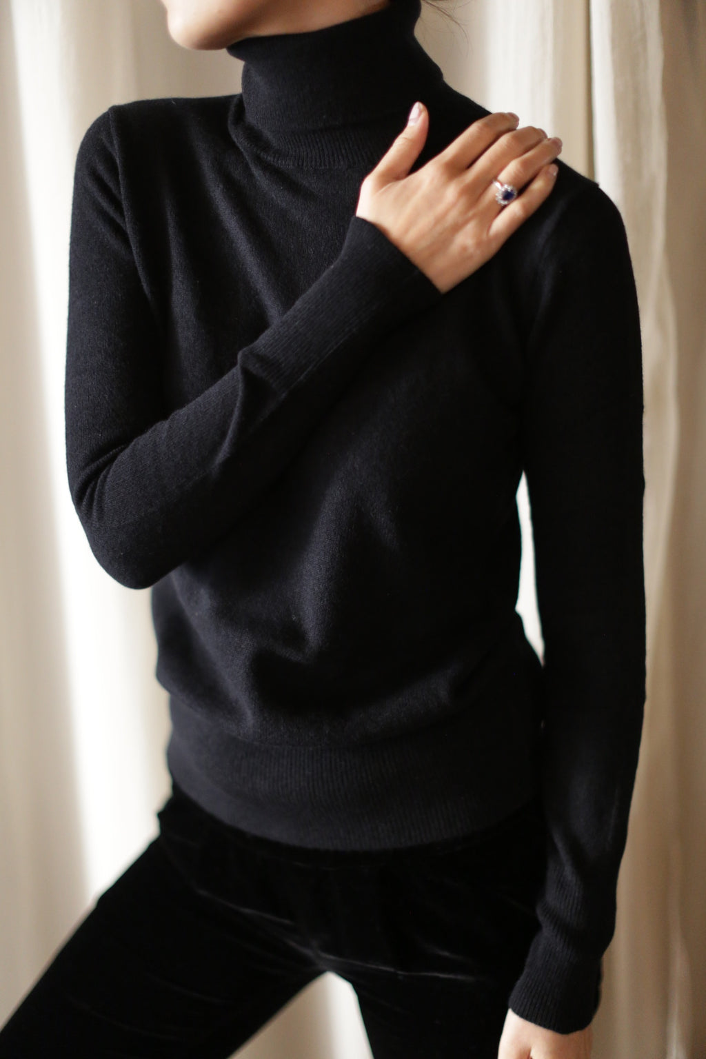 A person with an upright posture, wearing a long-sleeved black Cashmere Turtleneck and black pants, touches their right shoulder with their left hand. The background features light-colored curtains. The individual is showcasing a ring on their hand washable left hand garment.