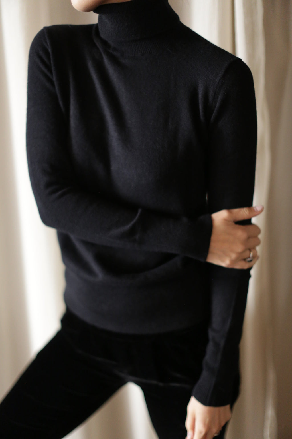 A person wearing a hand washable black Cashmere Turtleneck and black pants is standing in front of light-colored curtains. They have their right arm crossed over their body and are holding their left arm just above the elbow. Their face is not visible.
