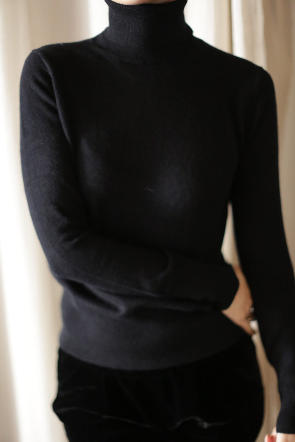 A person wearing a black Cashmere Turtleneck and dark pants is standing with their arms crossed in front of a cream-colored curtain. The image is cropped to show only the upper body.