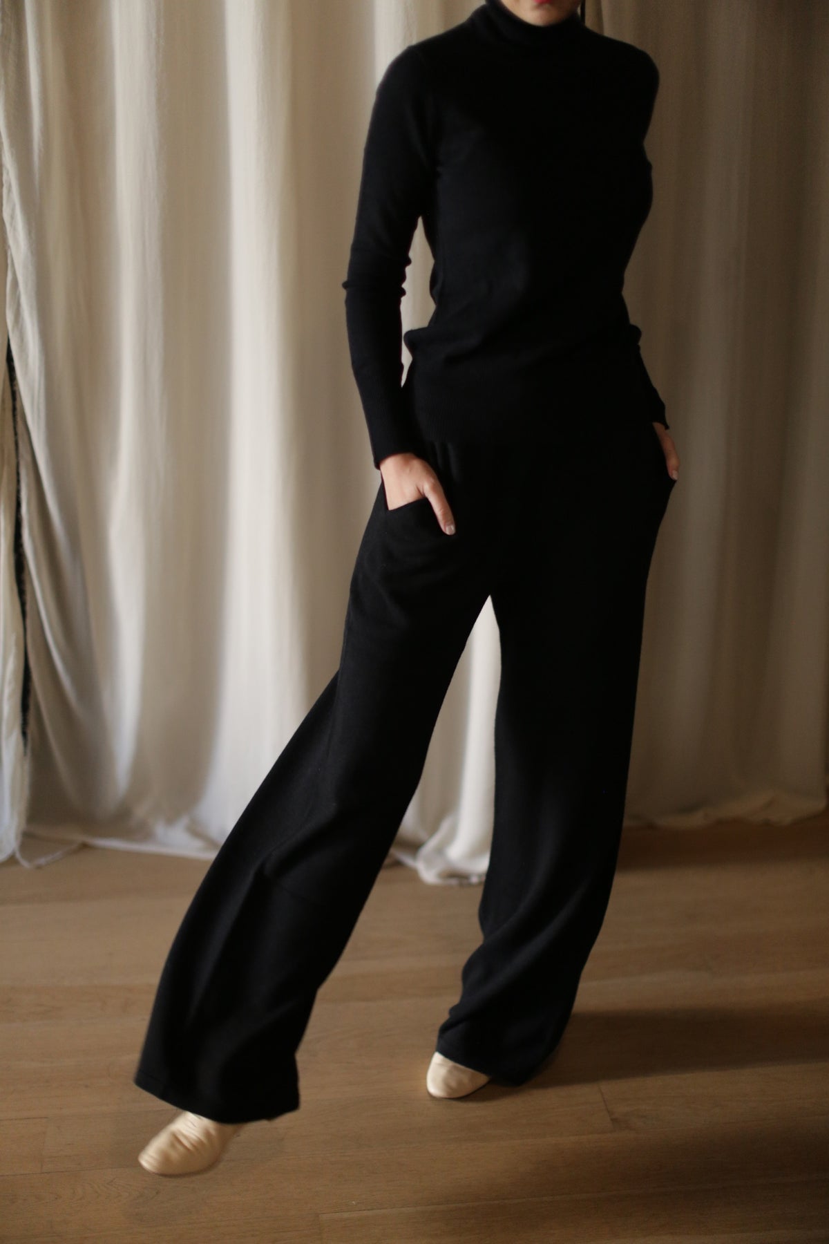 A person stands indoors on a wooden floor wearing Cashmere Wide Leg Pants with an elasticated waistband and pockets, paired with a matching black long-sleeve turtleneck. Their face is not visible; light curtains hang in the background.