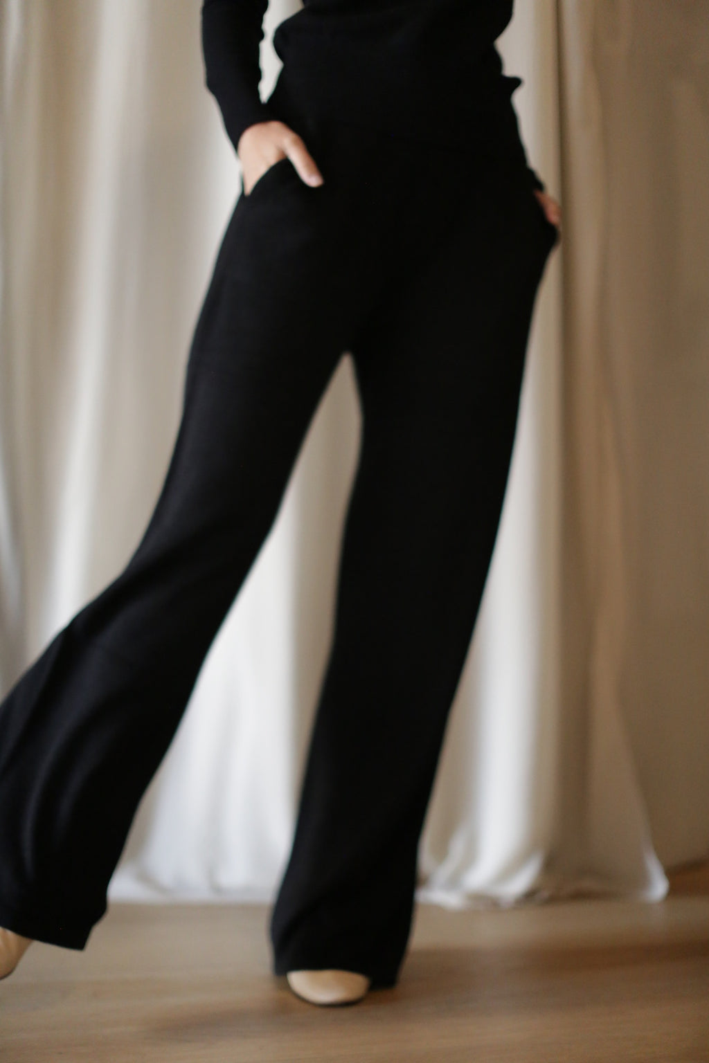 A person wears the Cashmere Wide Leg Pant with an elasticated waistband and a long-sleeve top, standing hands in pockets before cream-colored curtains, their face out of frame.