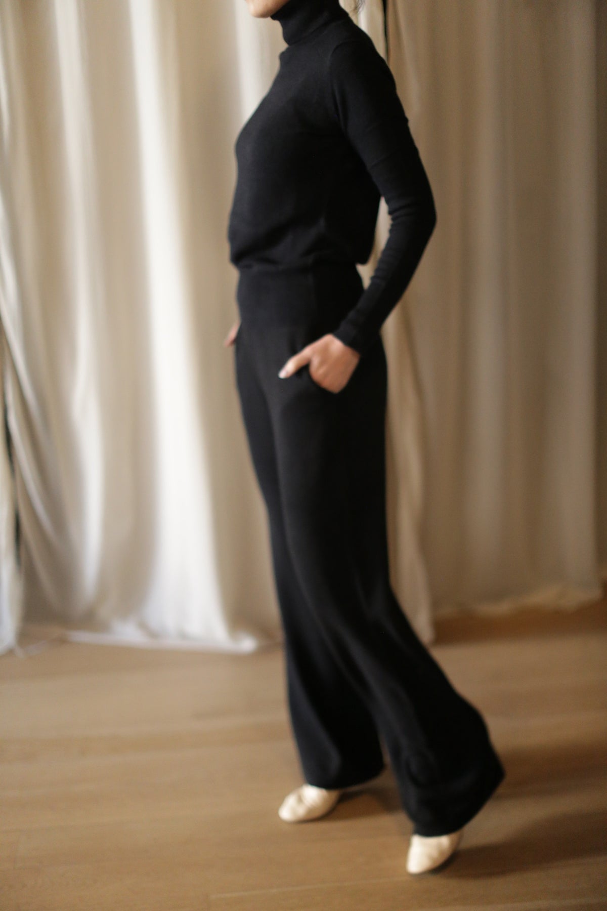 A person stands indoors, hands in pockets, wearing the Cashmere Wide Leg Pant in black with an elasticated waistband and a matching turtleneck. The wooden floor and light curtains are visible in the background; their face is not shown.