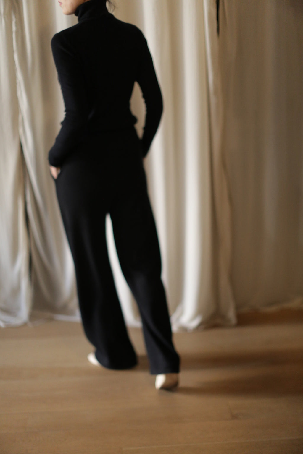 A person wears the Cashmere Wide Leg Pant with an elasticated waistband and a black long-sleeve top, standing indoors on a wooden floor before light-colored curtains, facing slightly away from the camera.
