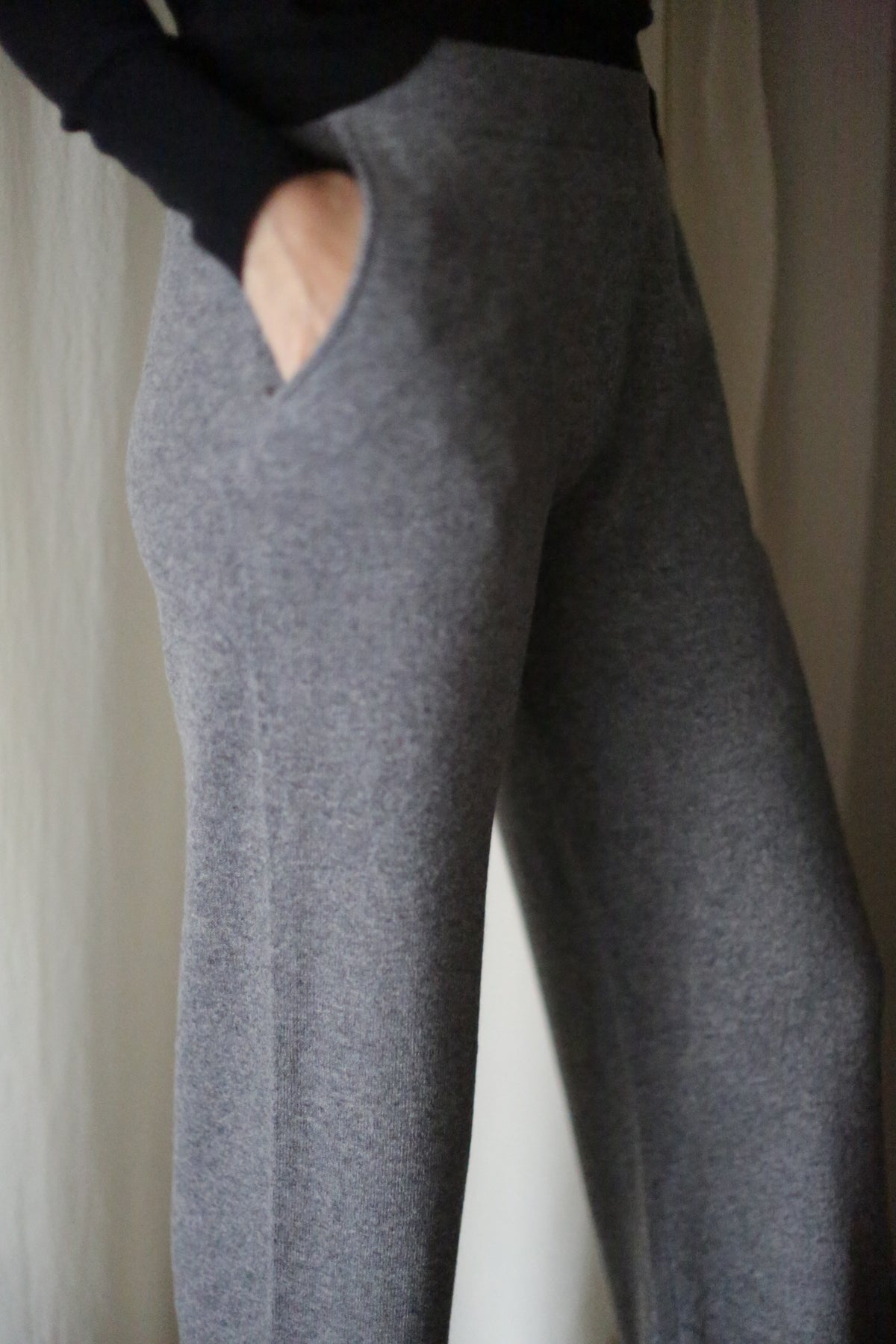 A person models the Cashmere Wide Leg Pant—gray, high-waisted, wide-leg with an elastic waistband and pocket—indoors against a light background, showing only the lower torso and legs.