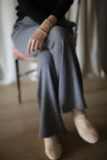 A person sits cross-legged on a chair, wearing the Cashmere Wide Leg Pant in gray with an elasticated waistband, paired with a black top, beige shoes, and bracelets. The background is softly blurred.