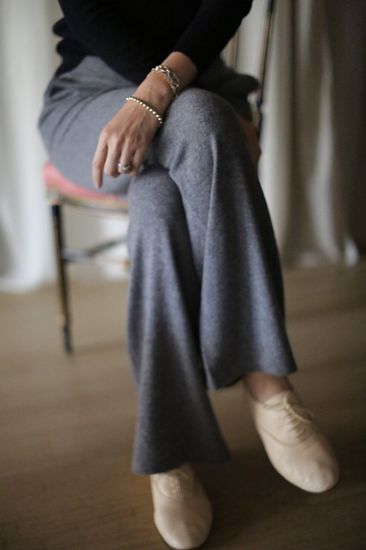A person sits cross-legged on a chair, wearing the Cashmere Wide Leg Pant in gray with an elasticated waistband, paired with a black top, beige shoes, and bracelets. The background is softly blurred.
