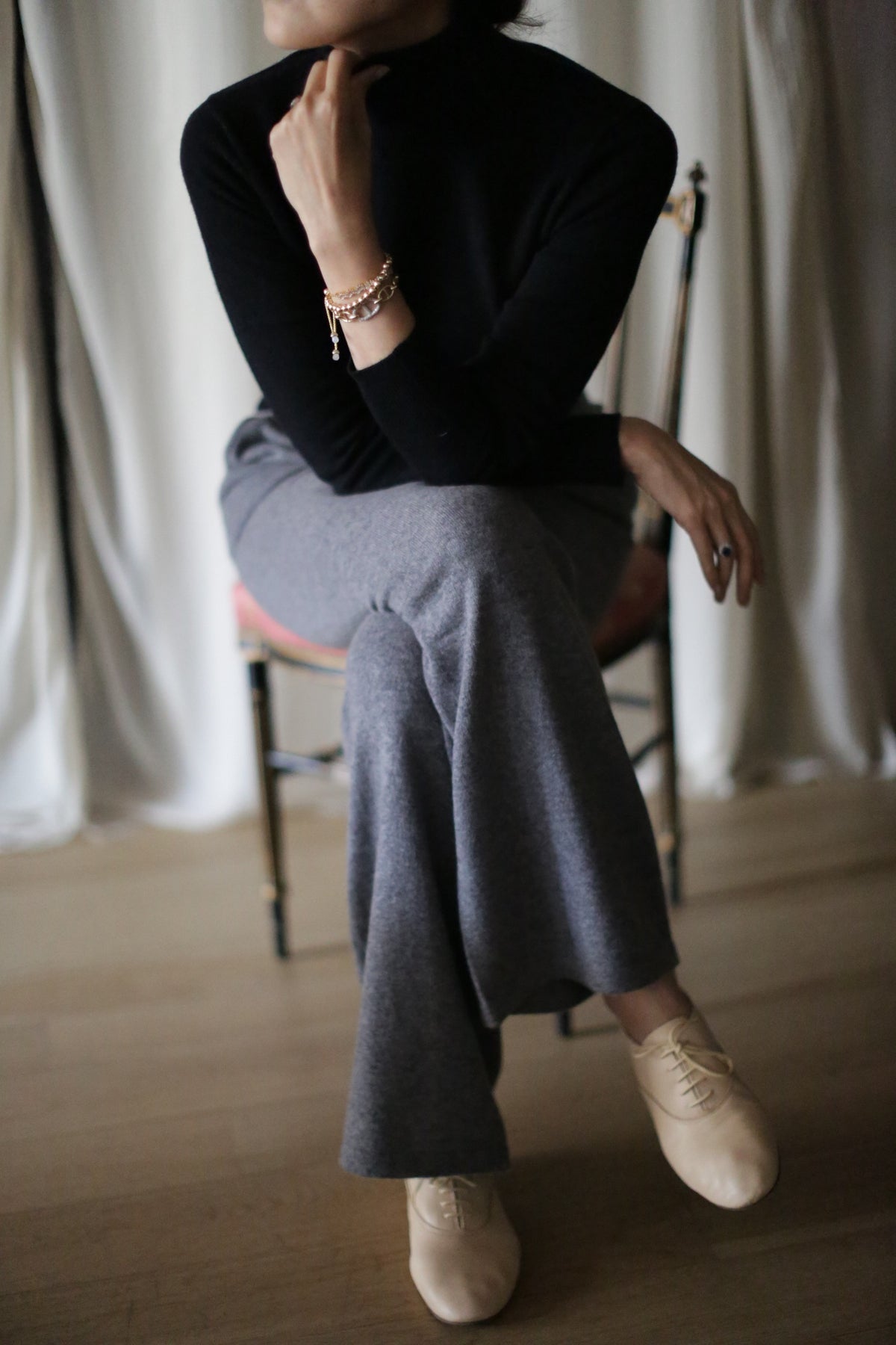 A person sits cross-legged on a chair, wearing Cashmere Wide Leg Pants in gray with an elasticated waistband, a black turtleneck, beige shoes, and bracelets. Their hand partially obscures their face. Pale curtains hang behind them.