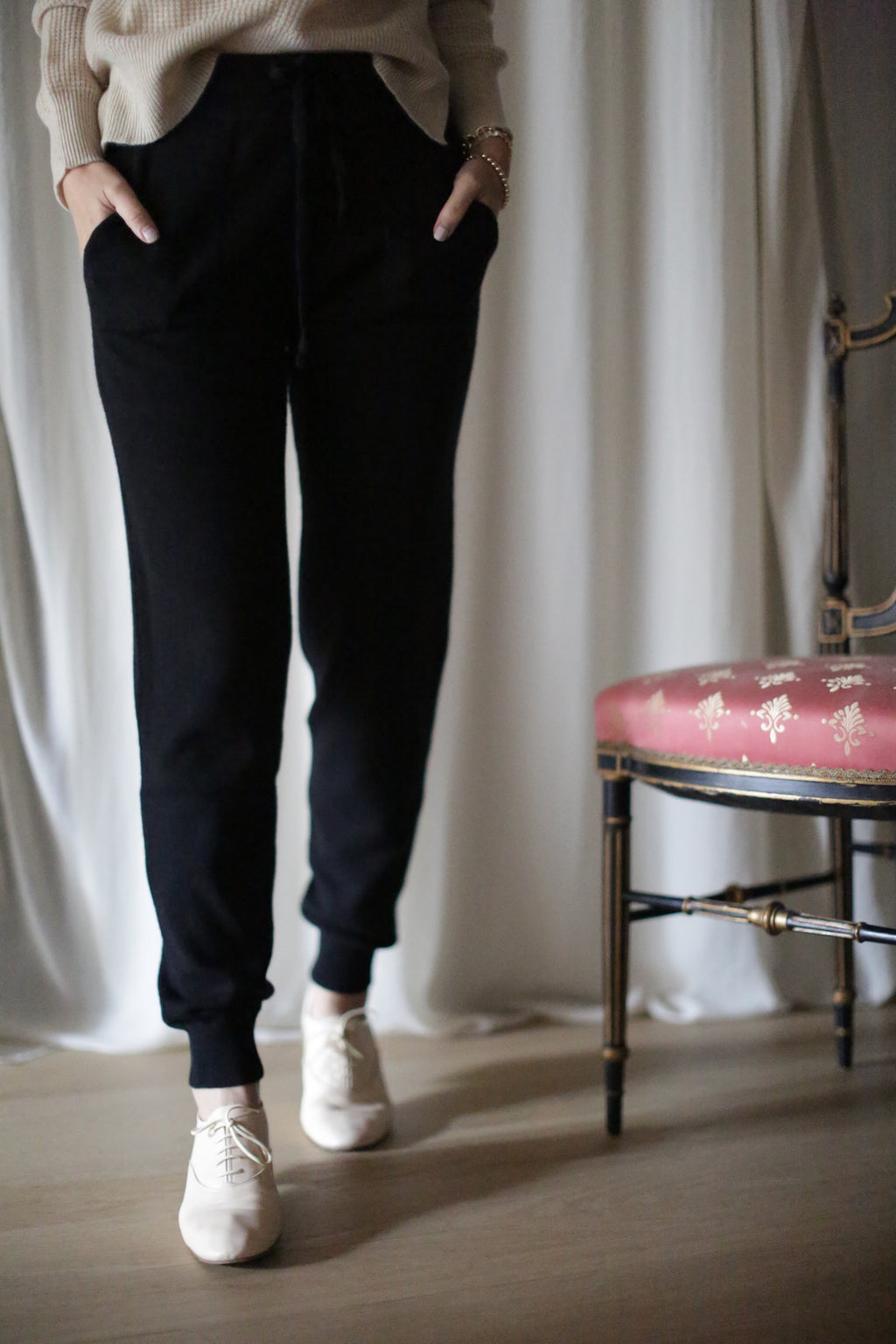 A person is indoors wearing black Cashmere Joggers with a drawstring and beige shoes, hands in pockets. A red chair with a gold floral pattern is partially visible on the right, set against white draped curtains.
