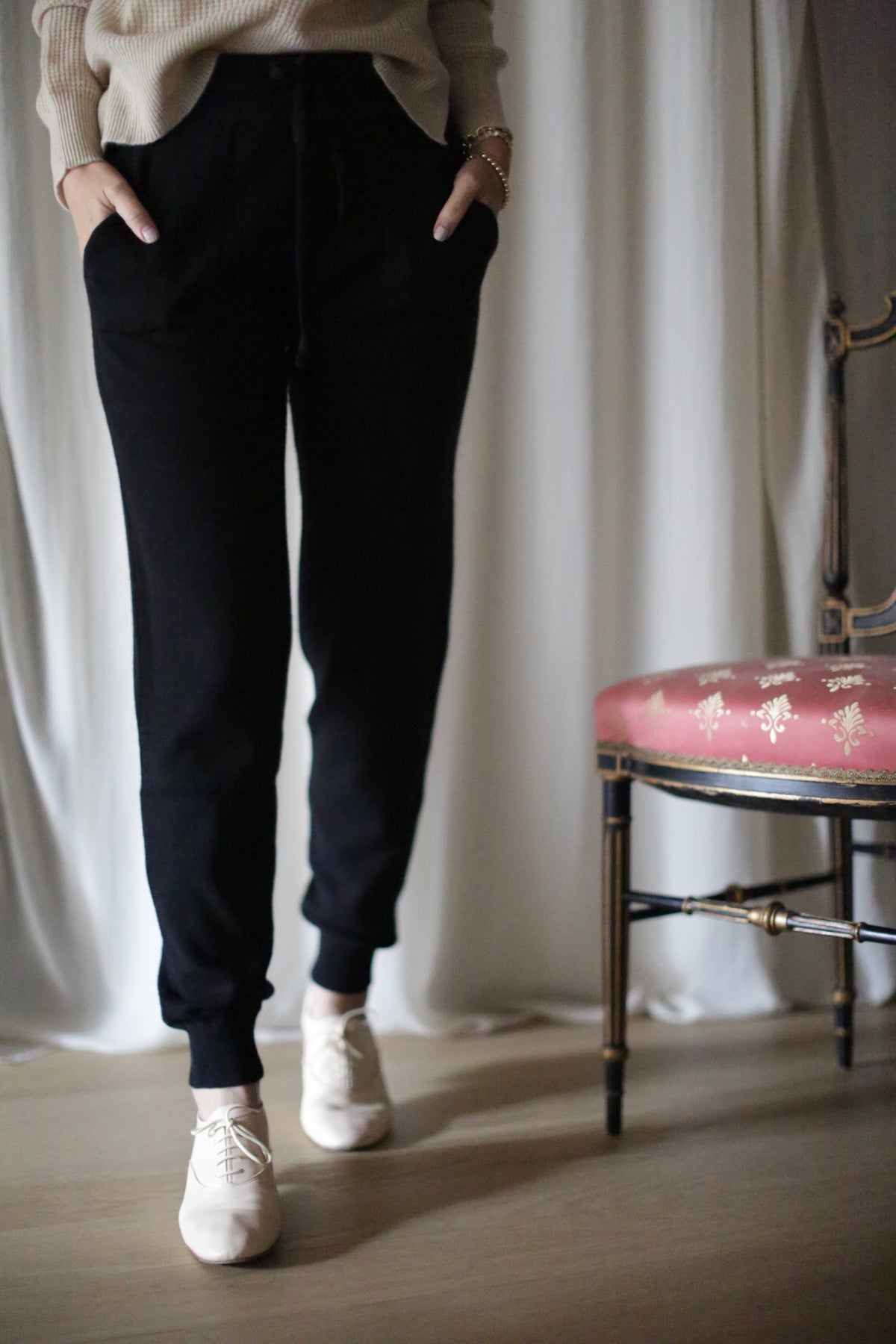 A person is indoors wearing black Cashmere Joggers with a drawstring and beige shoes, hands in pockets. A red chair with a gold floral pattern is partially visible on the right, set against white draped curtains.