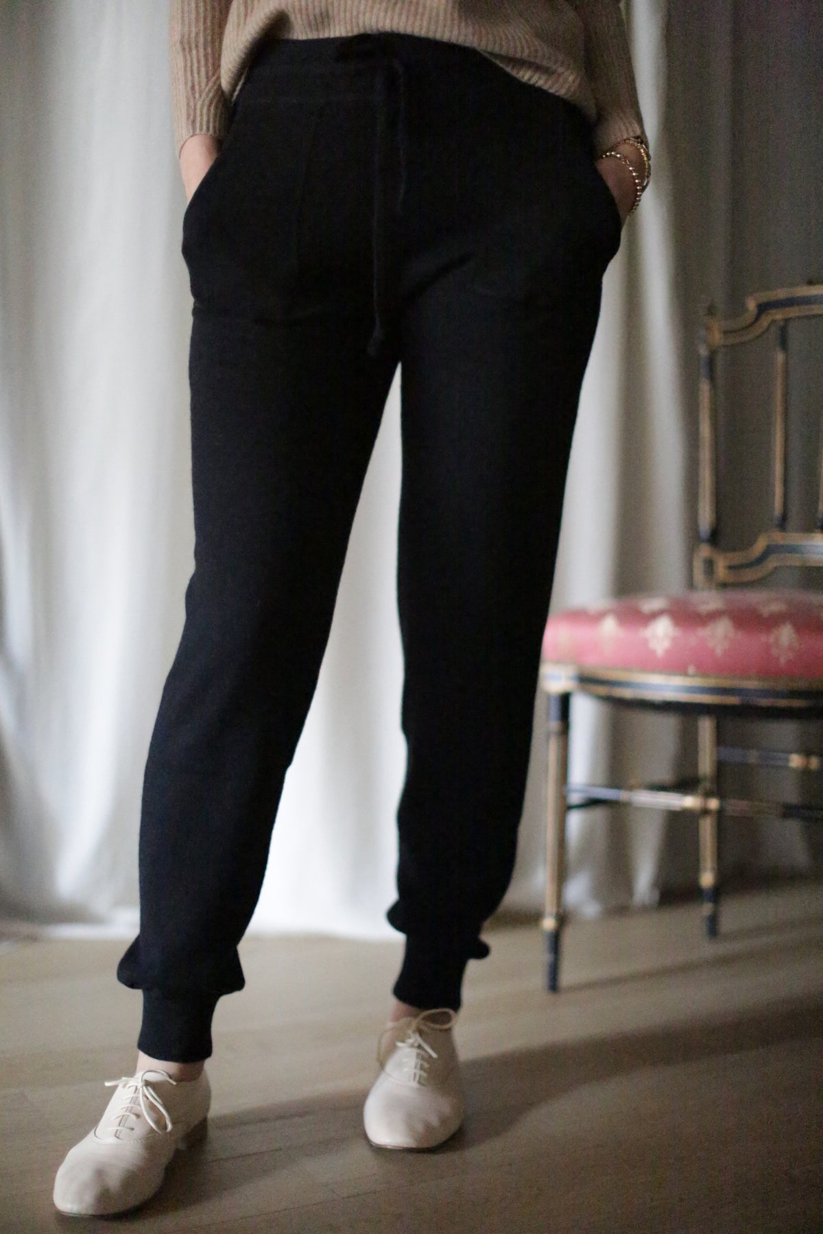 Wearing cashmere joggers and white sneakers, a person stands indoors with hands in pockets, partially visible from the waist down. A wooden chair with a pink cushion is seen in the background against a white curtain.