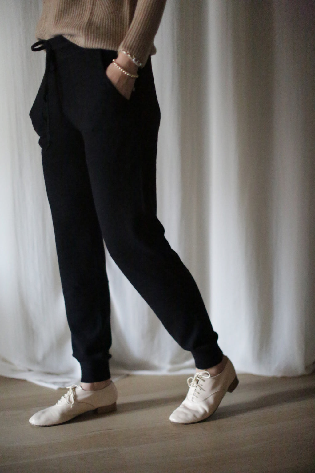 A person in a beige cashmere sweater and cashmere joggers with a drawstring stands with one hand in their pocket. They wear white lace-up shoes on a light wooden floor, against a white curtain backdrop.