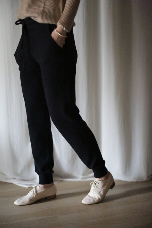 A person in a beige cashmere sweater and cashmere joggers with a drawstring stands with one hand in their pocket. They wear white lace-up shoes on a light wooden floor, against a white curtain backdrop.