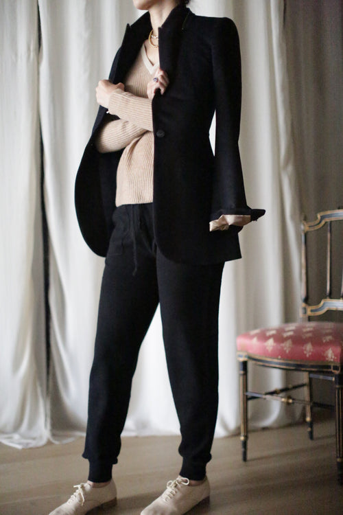A person stands before white curtains, wearing cashmere joggers with a black blazer and pants, sneakers, and a beige top. A pink patterned chair is partially visible to the right. The image emphasizes the textures and colors of the outfit rather than focusing on the person's face.