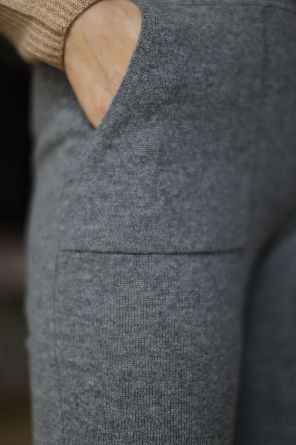A person casually tucks their hand into the pocket of Cashmere Joggers. The soft, textured gray knit pants feature a drawstring and evoke effortless elegance paired with a tan sweater, blending style with practicality.