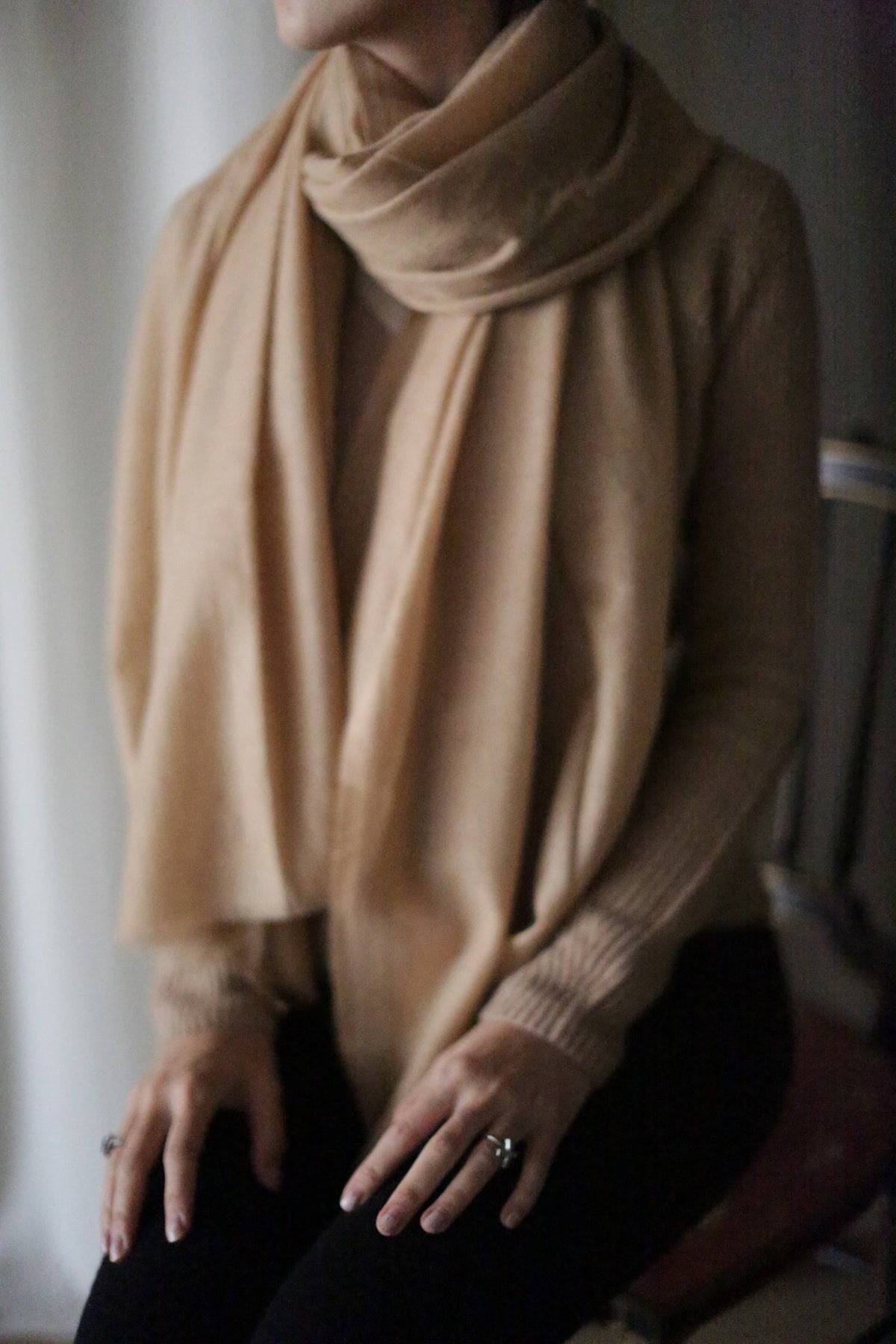 Seated indoors with a softly blurred background, a person is draped in a Tissue Weight Cashmere Scarf in Camel and a matching sweater. Their hands, resting on their lap, are adorned with silver rings. The person's face remains unseen in the image.