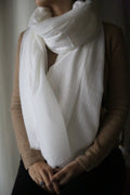 A person wearing a light brown sweater and a large, ivory Tissue Weight Cashmere Scarf wrapped around their neck. The person's hands are resting in their lap, with one hand holding the other. The background is softly blurred, focusing attention on the person and their attire.