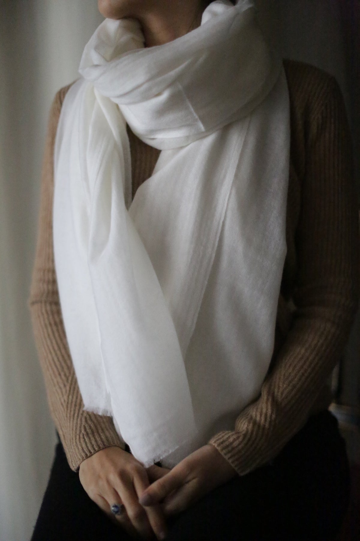 A person wearing a light brown sweater and a large, ivory Tissue Weight Cashmere Scarf wrapped around their neck. The person's hands are resting in their lap, with one hand holding the other. The background is softly blurred, focusing attention on the person and their attire.