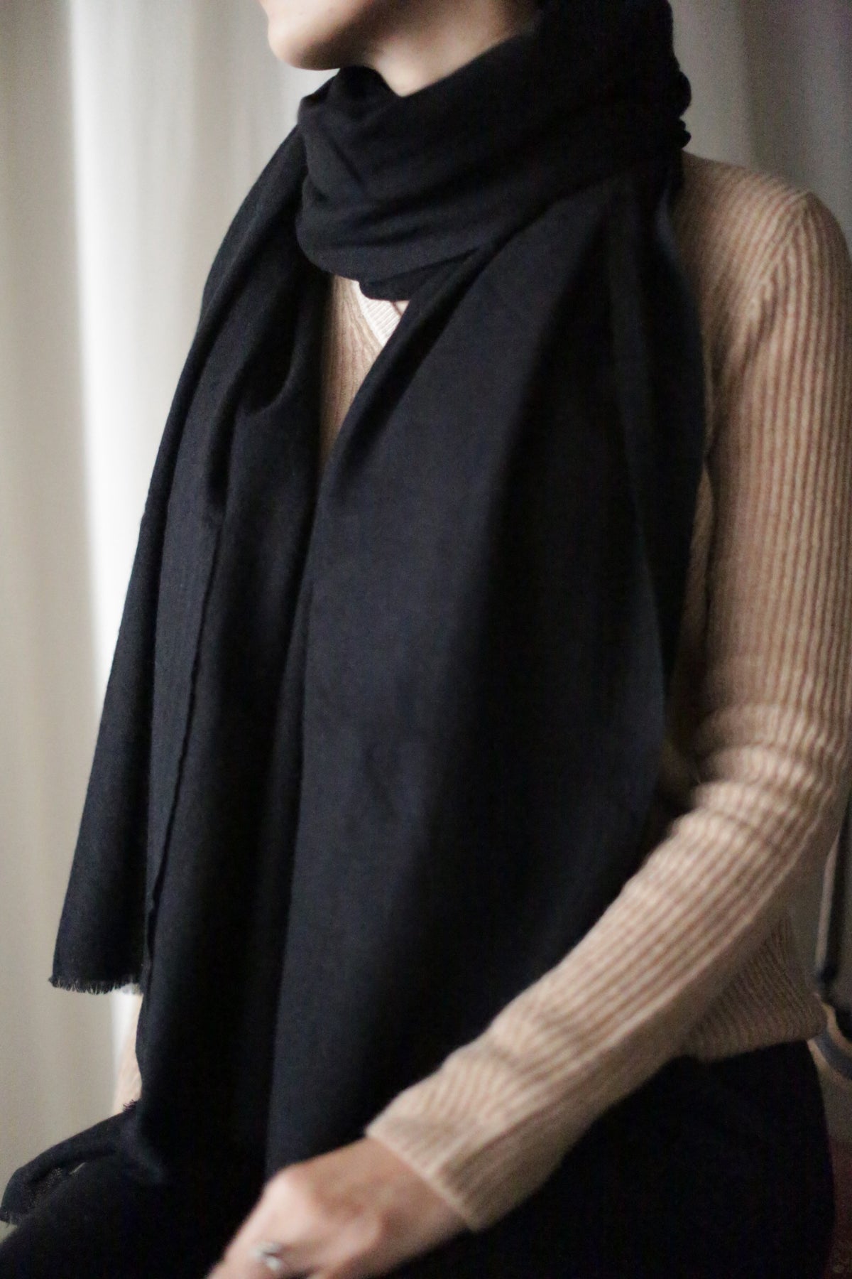 A person wearing a beige sweater and a large Tissue Weight Cashmere Scarf in black wrapped around their neck. The person is partially out of frame, with their face not visible. The background is softly lit with white drapes.