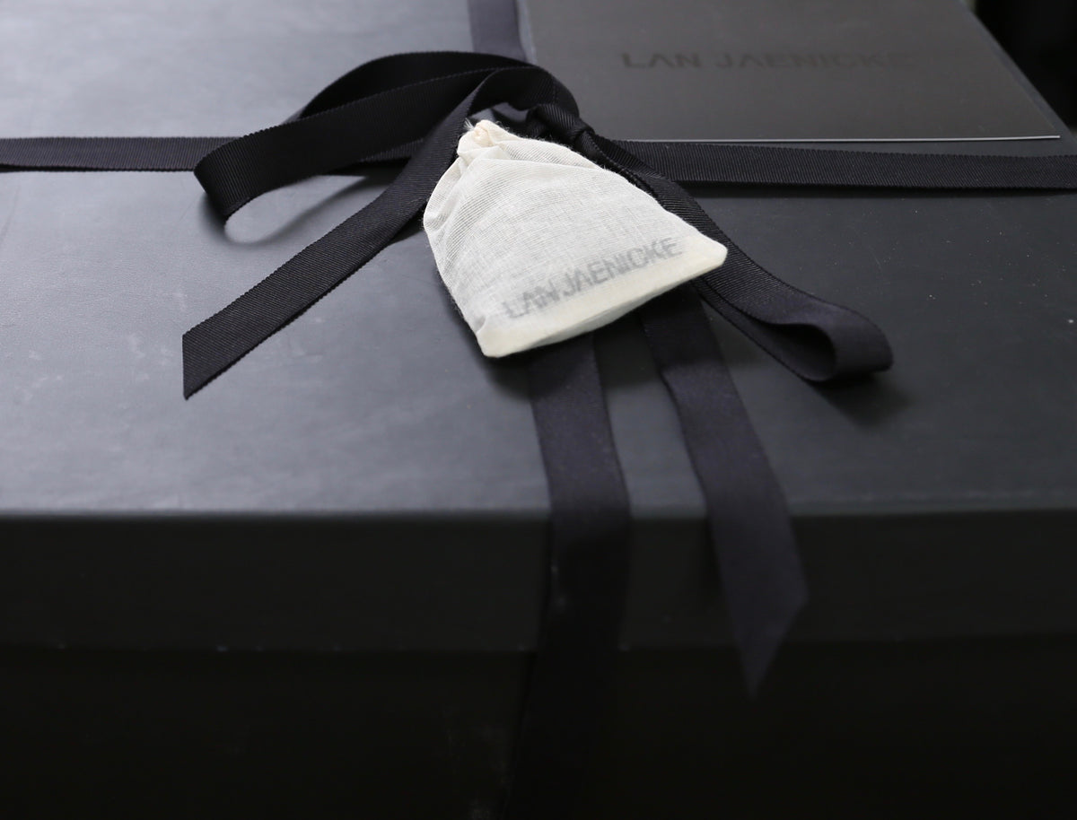 A small white drawstring pouch with the "LAN JAENICKE" text printed on it is tied to a black gift box with a black ribbon. The box, also placed in the background, features the same brand name, "LAN JAENICKE," printed on it. This elegant packaging holds a Gift Card. For more details or to browse in-store collections, visit lanjaenicke.com.
