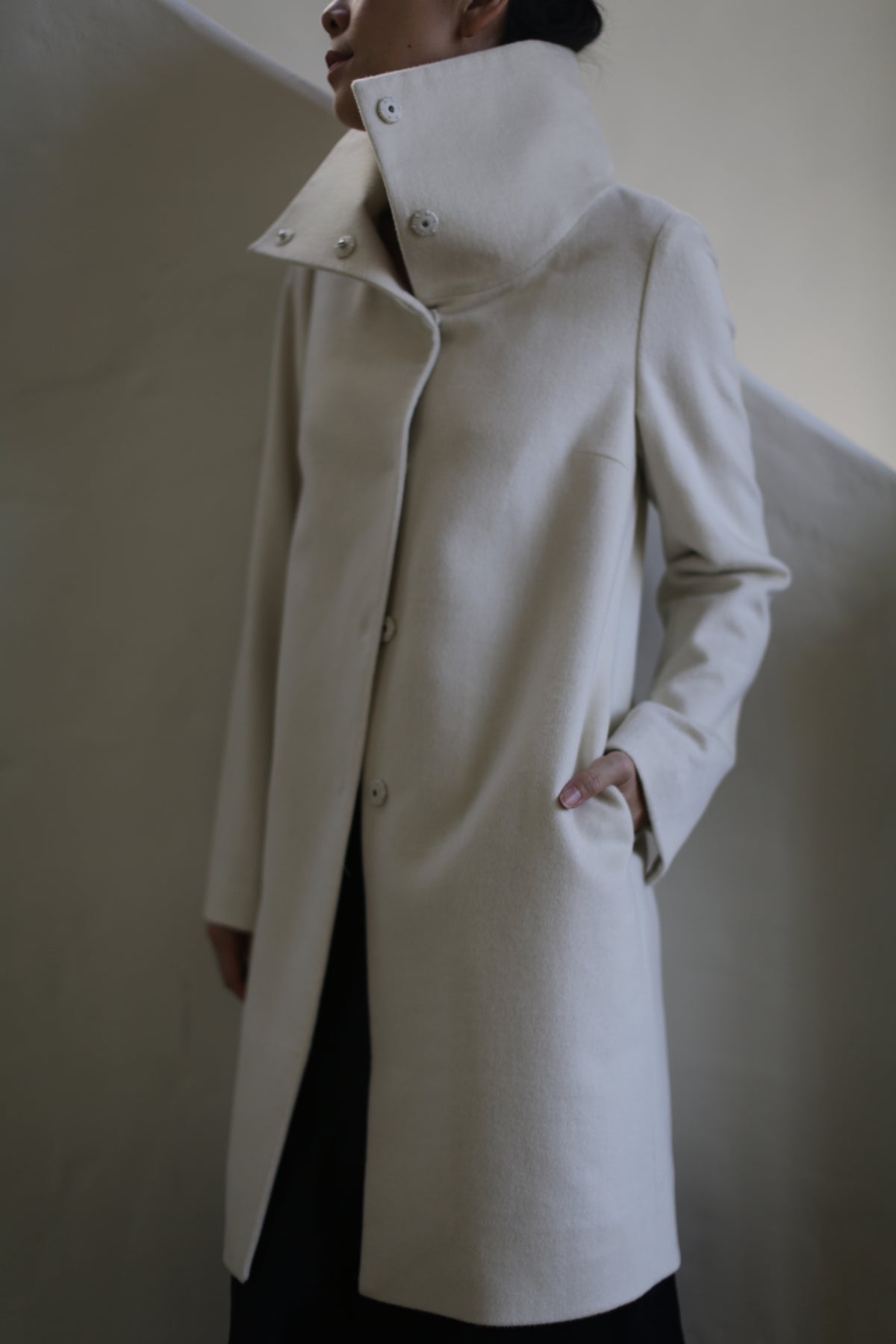 Dressed in the Stamford Long | Undyed, a tailored light beige cashmere coat with a high collar, the person stands against a minimalist neutral backdrop. The coat's button row and hand-in-pocket pose exude modern sophistication.