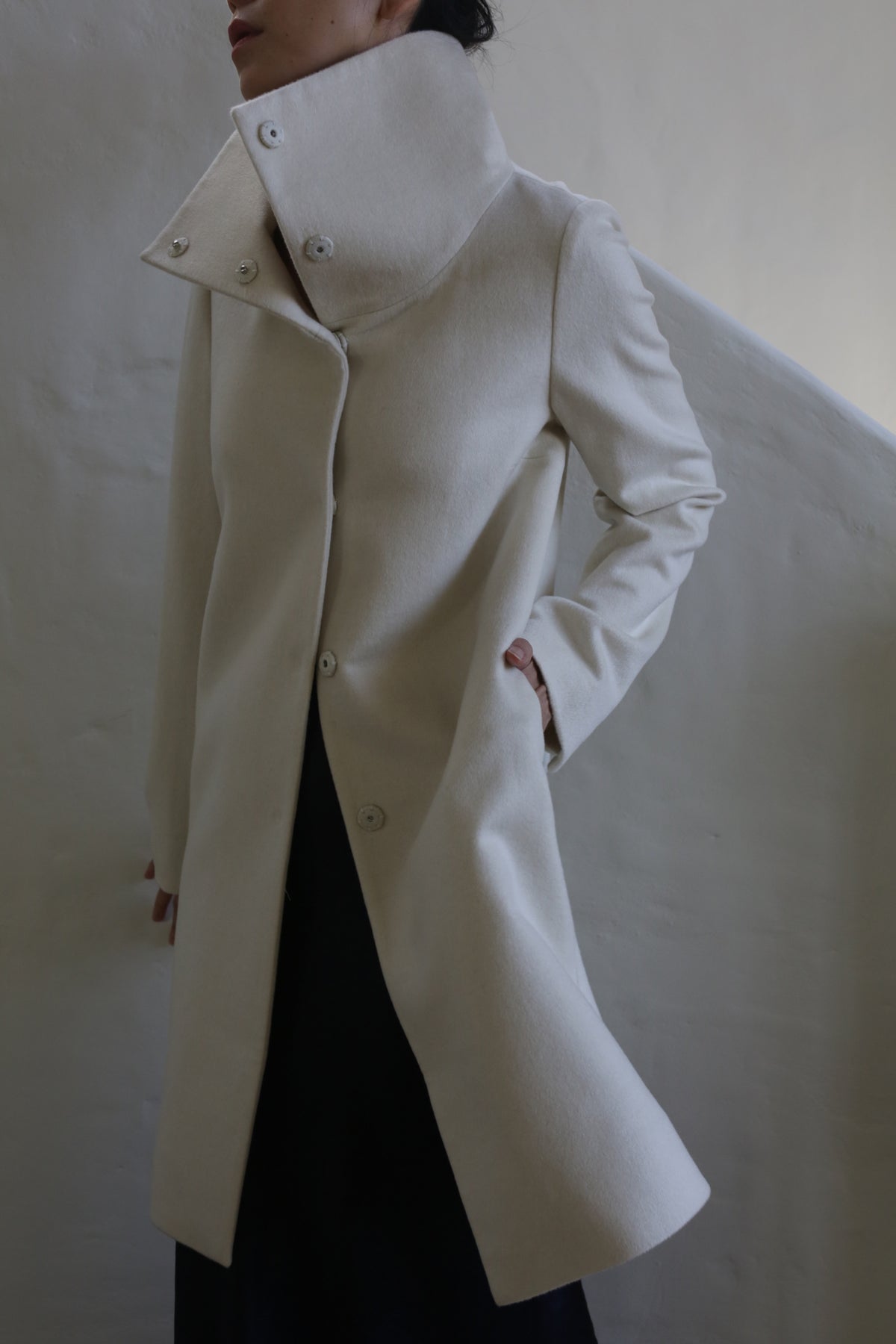 Wearing the Stamford Long | Undyed, a stylish cashmere coat with a high collar and large buttons, a person stands against a neutral background. The coat's modern, minimalist design is showcased as their hands rest casually in the pockets, with their face partially obscured.