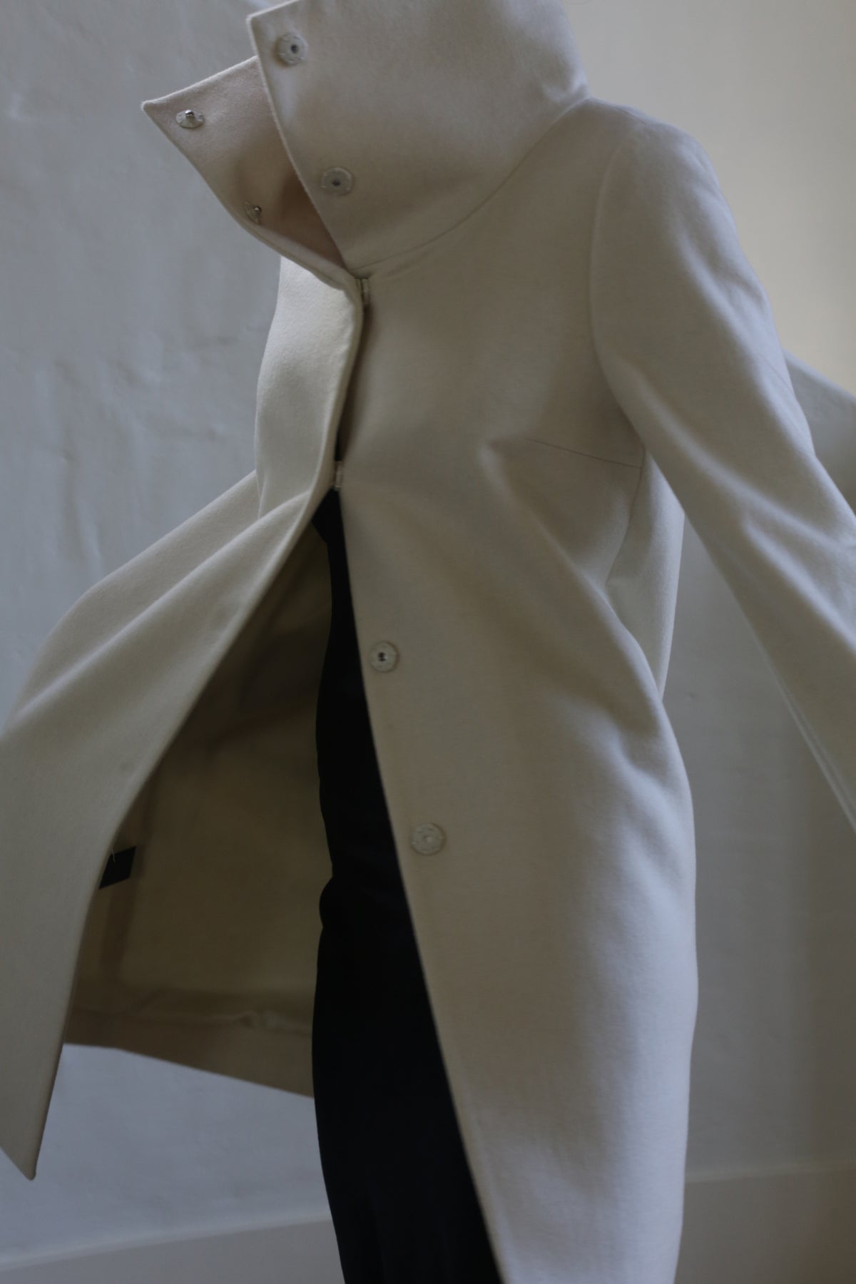 A person wears a Stamford Long | Undyed coat in cashmere featuring a high collar and large buttons. The coat is slightly open, exposing a dark outfit beneath, set against a plain, neutral-toned wall.