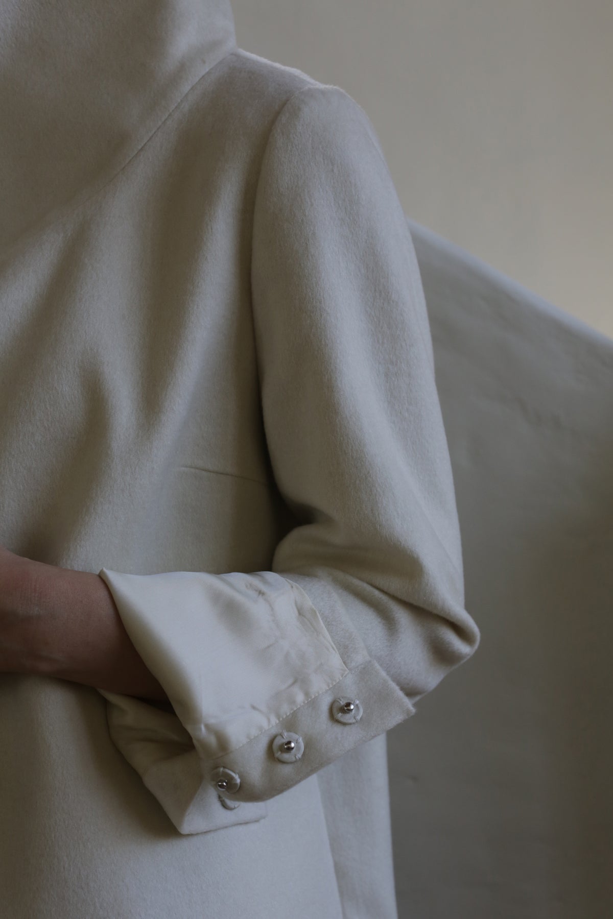 Close-up of a person wearing the Stamford II | Undyed coat with their arm partially visible. The high collar coat features cuffs adorned with neatly folded fabric and three decorative, shiny metallic buttons. The background is softly lit and out of focus.