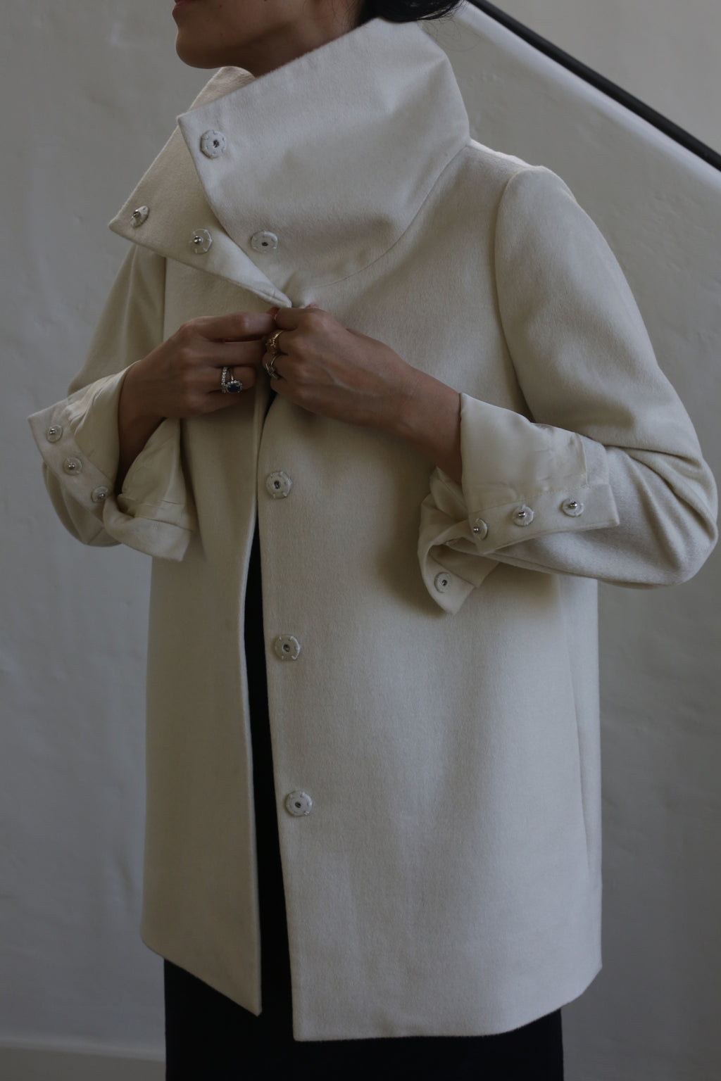 A person wearing the Stamford II | Undyed, a high-collared coat crafted from woven cashmere, adjusts the button-secured cuffs. The scene is set against a simple, light-colored wall and staircase, with the individual's face not visible.